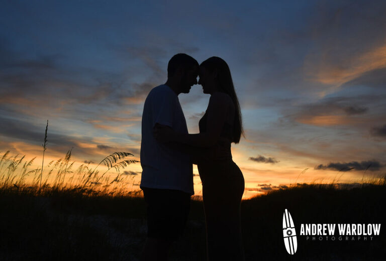 A happy couple is silhouetted against a beautiful Panama City Beach sunset after getting engaged during a beach proposal photo session. 