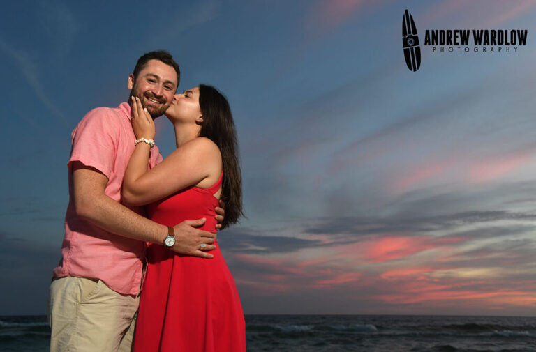 A woman kisses a man on the cheek during sunset after a beach proposal photo session in Panama City Beach, Florida.