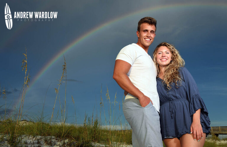 A couple poses for a photo with a rainbow before getting engaged at St. Andrews State Park in Panama City Beach, Florida. 