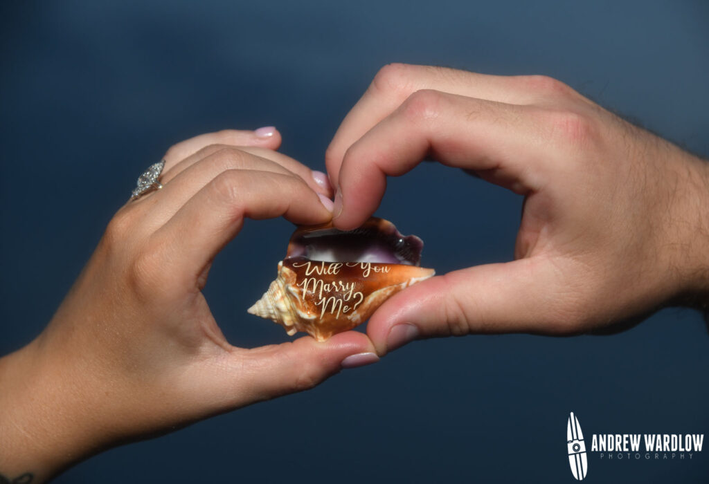 A man and a woman hold a shell that says, "Will you Marry Me?" during a beach proposal photo session in Panama City Beach, Florida.